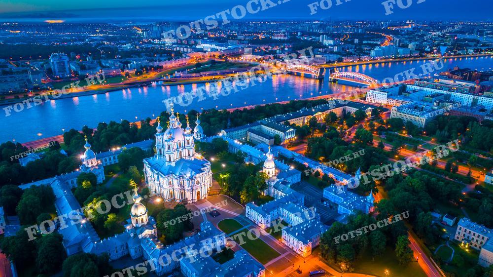 Этот снимок показывает Смольный собор в Санкт-Петербурге, сделанный с высоты в вечернее время. Яркие огни собора контрастируют с темнеющим небом, а его белоснежные стены и голубые купола явно выделяются в городском пейзаже. На заднем плане простирается вид на реку Неву и город с освещёнными улицами и мостами. Это изображение идеально подойдёт для публикаций о Санкт-Петербурге, туристических проспектов и культурных обзоров.